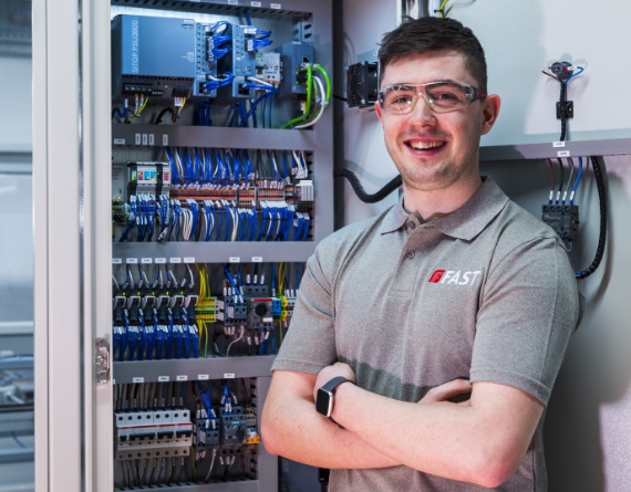 A photo of a FAST employee wearing safety goggles and smiling to the camera. They are standing in front of a electrical DC system cabinet.